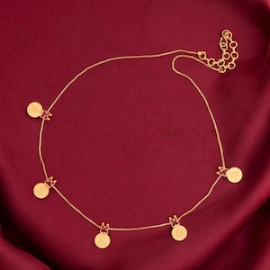 Puede incluir: Un collar dorado con cinco colgantes circulares. Cada colgante está adornado con una pequeña gema roja y un diseño en forma de corona. El collar se muestra sobre un fondo rojo oscuro texturizado.
