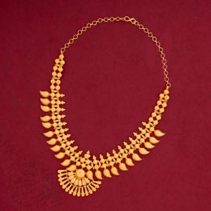 May include: A gold-colored necklace with a detailed design. The necklace features a series of leaf-shaped pendants and small, round beads. The necklace is set against a red background.