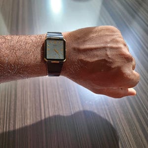 Puede incluir: Un reloj de pulsera rectangular dorado con esfera clara y correa negra. El reloj presenta números romanos y agujas doradas. El reloj se lleva en la muñeca.