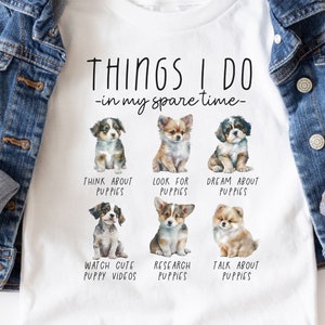 Op de afbeelding: Wit T-shirt met een blauw spijkerjasje. Het shirt heeft de tekst "THINGS I DO - in my spare time" en illustraties van puppy's. De activiteiten zijn: nadenken over puppy's, zoeken naar puppy's, dromen over puppy's, schattige puppyvideo's bekijken, puppy's onderzoeken en over puppy's praten.