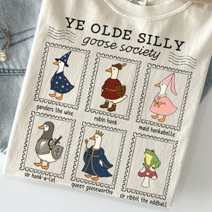 May include: Ivory-colored t-shirt with the text "YE OLDE SILLY goose society." The shirt features six cartoon goose characters in postage stamp-like frames. The geese are dressed in various costumes, including a wizard, a knight, and a queen.