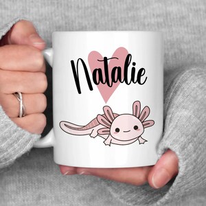 K&ouml;nnte beinhalten: Wei&szlig;e Keramiktasse mit dem Namen "Natalie" in schwarzer Schrift &uuml;ber einem rosa Herz und einer Cartoon-Axolotl-Illustration. Der Axolotl ist rosa mit schwarzen Augen und einem l&auml;chelnden Gesichtsausdruck. Die Tasse wird von einer Person gehalten.