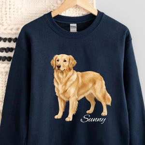 May include: Navy blue sweatshirt featuring a golden retriever illustration. The dog is depicted in a standing pose with a light brown coat. The word "Sunny" is written in a cursive font below the dog. The sweatshirt is hanging on a wooden hanger.