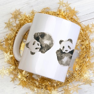 May include: White ceramic mug with a black and white illustration of two panda bears. The mug is surrounded by gold glitter stars.