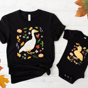 May include: Black t-shirt and onesie set featuring a white goose and yellow duck design surrounded by fall foliage. The goose and duck are surrounded by pumpkins, leaves, and hearts. A matching set for adults and babies.