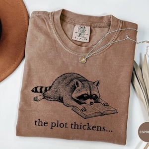 May include: A tan crew neck t-shirt with a black graphic print of a raccoon reading an open book. Below the raccoon, the text reads, the plot thickens... The shirt features a Comfort Colors brand label at the inner neckline with size S-CH.