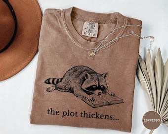 May include: A tan crew neck t-shirt with a black graphic print of a raccoon reading an open book. Below the raccoon, the text reads, the plot thickens... The shirt features a Comfort Colors brand label at the inner neckline with size S-CH.