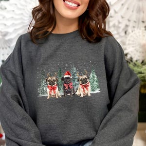 May include: A dark heather grey sweatshirt with a Christmas-themed design featuring three pugs wearing Santa hats and reindeer antlers. The pugs are standing in front of a snowy forest scene.
