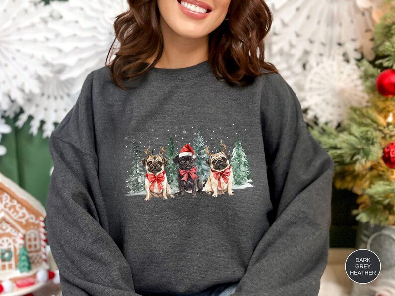 May include: A dark heather grey sweatshirt with a Christmas-themed design featuring three pugs wearing Santa hats and reindeer antlers. The pugs are standing in front of a snowy forest scene.