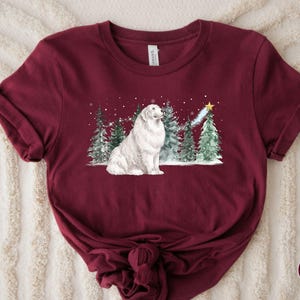 May include: Maroon t-shirt featuring a watercolor illustration of a white dog sitting in a snowy forest with Christmas trees. A shooting star with a gold star at the end is above the trees. The shirt has rolled-up sleeves and a knot at the bottom.