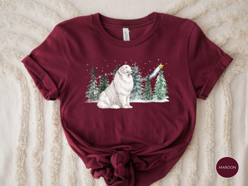 May include: Maroon t-shirt featuring a watercolor illustration of a white dog sitting in a snowy forest with Christmas trees. A shooting star with a gold star at the end is above the trees. The shirt has rolled-up sleeves and a knot at the bottom.