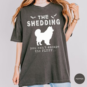 May include: Dark gray t-shirt with white text and a dog silhouette. The text reads "THE SHEDDING" above the dog and "you can't escape the FLUFF..." below. Two bat silhouettes are above the word "SHEDDING".