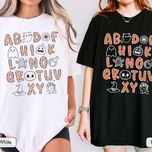 May include: A white t-shirt and a black t-shirt, each featuring a graphic print of the alphabet with orange and white checkered letters. The design includes small black line art illustrations of a ghost, pumpkin, mummy, skull, and witch hat.