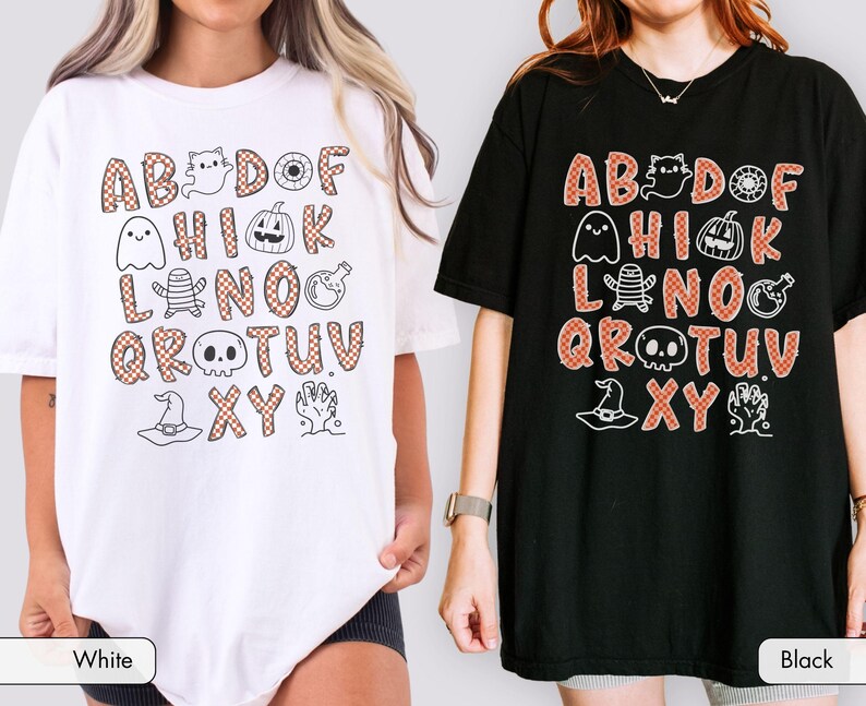 May include: A white t-shirt and a black t-shirt, each featuring a graphic print of the alphabet with orange and white checkered letters. The design includes small black line art illustrations of a ghost, pumpkin, mummy, skull, and witch hat.