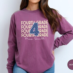 May include: A berry-colored long-sleeve sweatshirt featuring the text FOURTH GRADE repeated in a light cream color. A large navy blue number 4 with white polka dots is centered over the text. Below, the name Mrs. Smith is printed in white script.