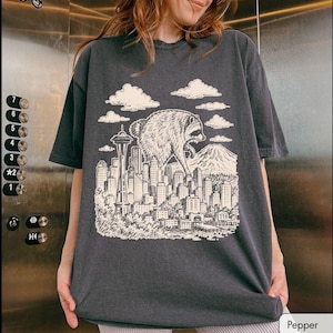 Pu&ograve; includere: Una t-shirt a maniche corte grigio antracite con una stampa grafica color crema raffigurante un procione gigante che sovrasta lo skyline di una citt&agrave;. L'illustrazione include lo Space Needle, vari grattacieli, una montagna sullo sfondo e nuvole.