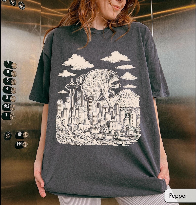 Pu&ograve; includere: Una t-shirt a maniche corte grigio antracite con una stampa grafica color crema raffigurante un procione gigante che sovrasta lo skyline di una citt&agrave;. L'illustrazione include lo Space Needle, vari grattacieli, una montagna sullo sfondo e nuvole.