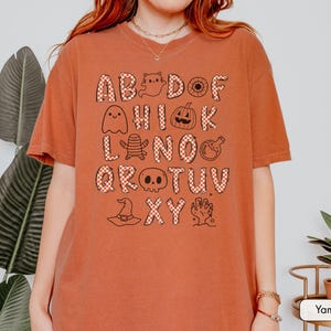 May include: A burnt orange short-sleeve t-shirt featuring a graphic print of the alphabet. Each letter is filled with a red and white checkered pattern, interspersed with black line-art illustrations of ghosts, pumpkins, skulls, and other spooky icons.