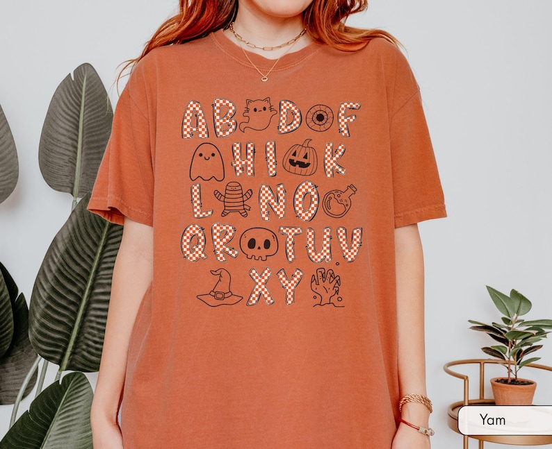 May include: A burnt orange short-sleeve t-shirt featuring a graphic print of the alphabet. Each letter is filled with a red and white checkered pattern, interspersed with black line-art illustrations of ghosts, pumpkins, skulls, and other spooky icons.