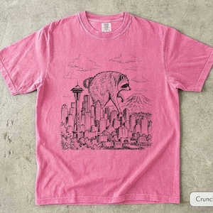 Pu&ograve; includere: Una t-shirt rosa a maniche corte con una grafica in stile inchiostro nero raffigurante un procione gigante che cammina sopra lo skyline di una citt&agrave;, inclusi lo Space Needle e una montagna sullo sfondo. La maglia ha un girocollo e un'etichetta.