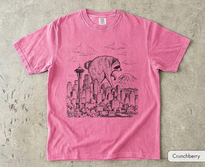 Pu&ograve; includere: Una t-shirt rosa a maniche corte con una grafica in stile inchiostro nero raffigurante un procione gigante che cammina sopra lo skyline di una citt&agrave;, inclusi lo Space Needle e una montagna sullo sfondo. La maglia ha un girocollo e un'etichetta.