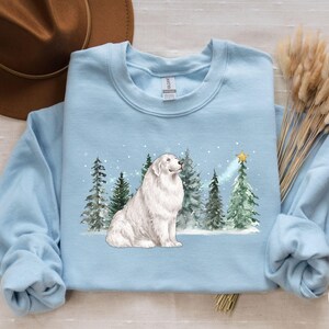 May include: Light blue sweatshirt featuring a white dog sitting in front of evergreen trees and a star. The dog has fluffy white fur. The trees are green and snow is falling. The words "LIGHT BLUE" are in a circle on the bottom right.