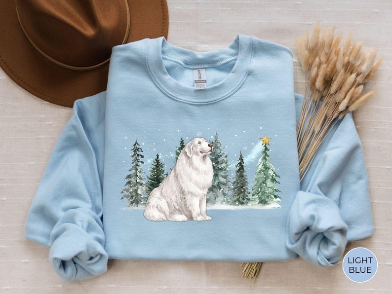 May include: Light blue sweatshirt featuring a white dog sitting in front of evergreen trees and a star. The dog has fluffy white fur. The trees are green and snow is falling. The words "LIGHT BLUE" are in a circle on the bottom right.