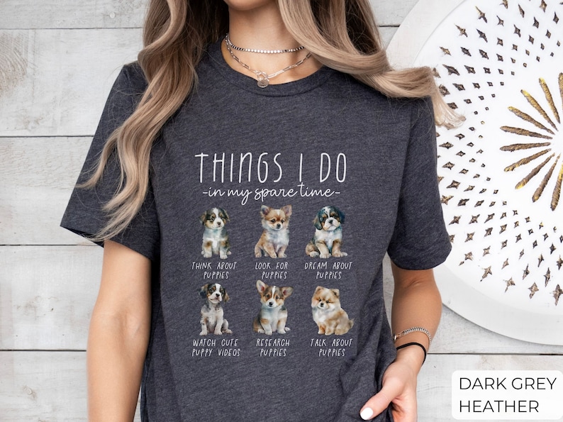 Op de afbeelding: Donkergrijze heather t-shirt met de tekst "THINGS I DO - in my spare time" en afbeeldingen van puppy's. Het shirt vermeldt activiteiten zoals "Think About Puppies" en "Watch Cute Puppy Videos".