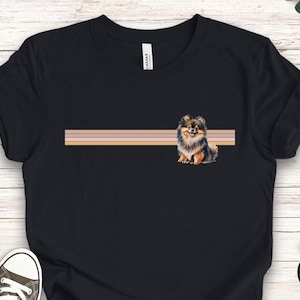 Puede incluir: Camiseta negra con una ilustraci&oacute;n de un perro pomerania marr&oacute;n y negro. El perro est&aacute; sentado frente a un dise&ntilde;o a rayas rosa, melocot&oacute;n y beige.