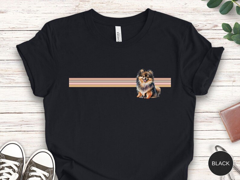 Puede incluir: Camiseta negra con una ilustraci&oacute;n de un perro pomerania marr&oacute;n y negro. El perro est&aacute; sentado frente a un dise&ntilde;o a rayas rosa, melocot&oacute;n y beige.