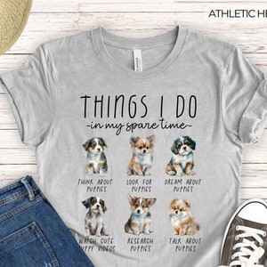Op de afbeelding: Athletic heather grijs t-shirt met de tekst "THINGS I DO - in my spare time-" en aquarel illustraties van puppy's. Het shirt zegt ook "THINK ABOUT PUPPIES", "LOOK FOR PUPPIES", "DREAM ABOUT PUPPIES", "WATCH CUTE PUPPY VIDEOS", "RESEARCH PUPPIES" en "TALK ABOUT PUPPIES".