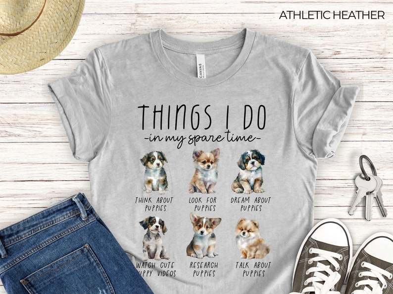 Op de afbeelding: Athletic heather grijs t-shirt met de tekst "THINGS I DO - in my spare time-" en aquarel illustraties van puppy's. Het shirt zegt ook "THINK ABOUT PUPPIES", "LOOK FOR PUPPIES", "DREAM ABOUT PUPPIES", "WATCH CUTE PUPPY VIDEOS", "RESEARCH PUPPIES" en "TALK ABOUT PUPPIES".