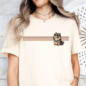 Puede incluir: Una camiseta blanca con un dise&ntilde;o de rayas marrones, negras y crema y una peque&ntilde;a imagen de un perro pomerania marr&oacute;n y negro.