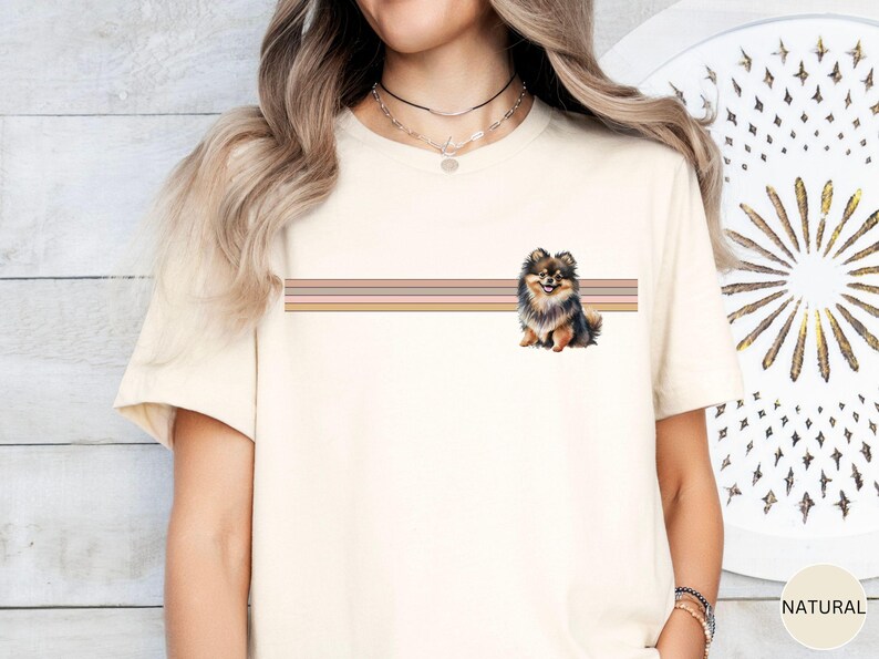 Puede incluir: Una camiseta blanca con un dise&ntilde;o de rayas marrones, negras y crema y una peque&ntilde;a imagen de un perro pomerania marr&oacute;n y negro.