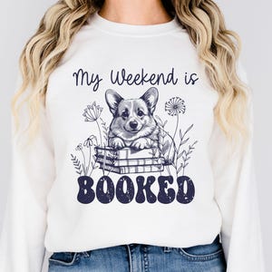 Puede incluir: Sudadera blanca con un gr&aacute;fico de un perro corgi sentado en una pila de libros. El texto "My Weekend is Booked" est&aacute; impreso debajo del perro.
