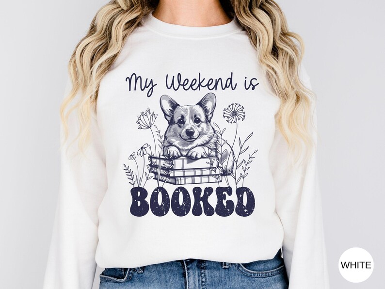 Puede incluir: Sudadera blanca con un gr&aacute;fico de un perro corgi sentado en una pila de libros. El texto "My Weekend is Booked" est&aacute; impreso debajo del perro.