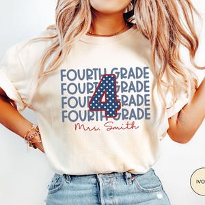 May include: An ivory short-sleeve t-shirt featuring the text "FOURTH GRADE" repeated four times in blue, with a large central number "4" in blue with white polka dots and a red outline, and the name "Mrs. Smith" in red cursive script below.