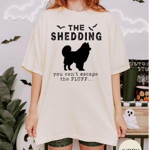 May include: Ivory-colored t-shirt with the text "THE SHEDDING" above a black silhouette of a dog. Below the dog, the text reads "you can't escape the FLUFF...". The shirt has short sleeves and a relaxed fit, perfect for casual wear.