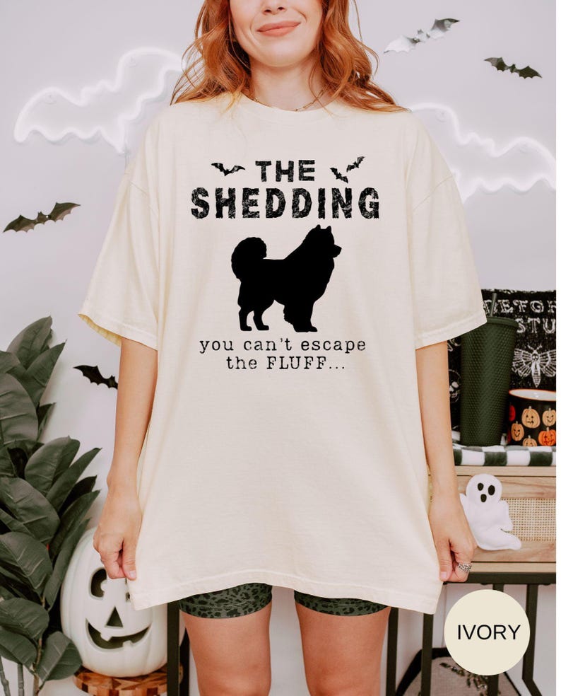 May include: Ivory-colored t-shirt with the text "THE SHEDDING" above a black silhouette of a dog. Below the dog, the text reads "you can't escape the FLUFF...". The shirt has short sleeves and a relaxed fit, perfect for casual wear.
