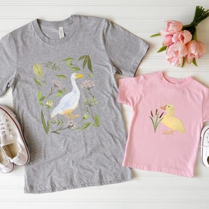 May include: Two t-shirts with a duck and goose design. The adult shirt is grey with a white goose and green foliage. The child's shirt is pink with a yellow duck and green reeds.