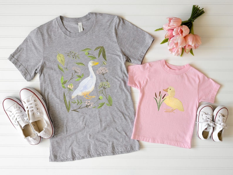May include: Two t-shirts with a duck and goose design. The adult shirt is grey with a white goose and green foliage. The child's shirt is pink with a yellow duck and green reeds.
