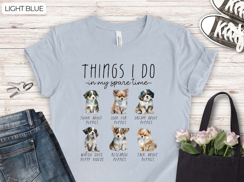 Op de afbeelding: Lichtblauw T-shirt met de tekst "THINGS I DO in my spare time" en illustraties van puppy's. Het shirt toont zes verschillende puppyrassen met bijschriften als "Think About Puppies" en "Watch Cute Puppy Videos."