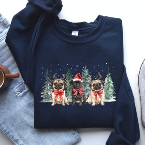 May include: A navy blue sweatshirt with a Christmas-themed design featuring three pugs wearing Santa hats and reindeer antlers. The pugs are surrounded by pine trees and snowflakes.