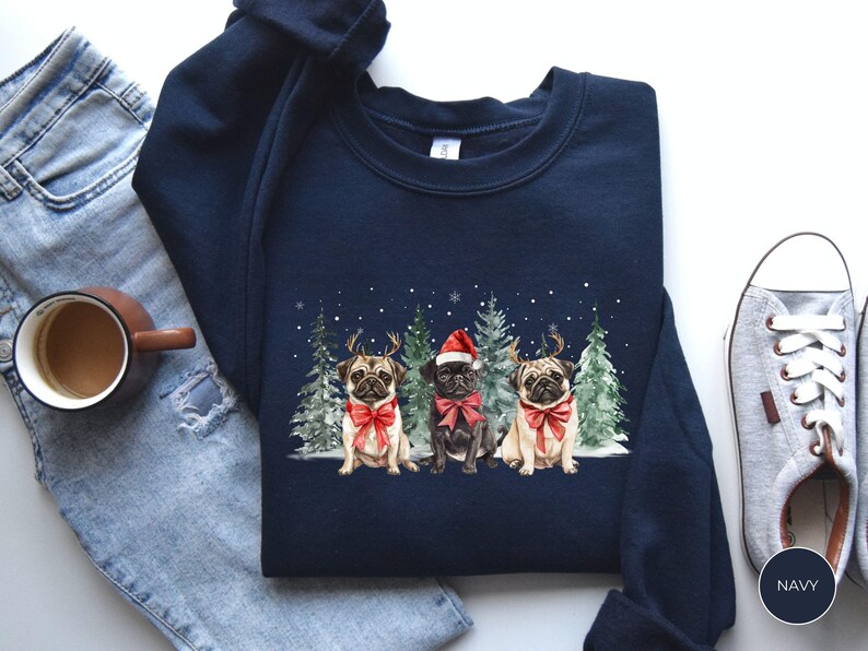May include: A navy blue sweatshirt with a Christmas-themed design featuring three pugs wearing Santa hats and reindeer antlers. The pugs are surrounded by pine trees and snowflakes.