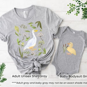 May include: A gray adult unisex shirt and a gray baby bodysuit. The shirt features a white goose with green foliage and flowers. The bodysuit features a yellow duckling with green reeds. The text "Adult gray and baby gray may not be an exact shade match" is printed below the images.