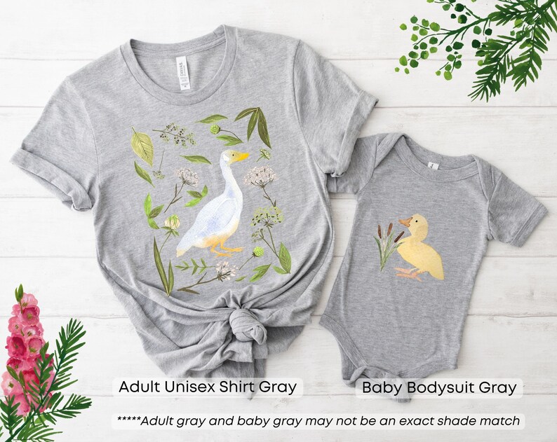 May include: A gray adult unisex shirt and a gray baby bodysuit. The shirt features a white goose with green foliage and flowers. The bodysuit features a yellow duckling with green reeds. The text "Adult gray and baby gray may not be an exact shade match" is printed below the images.