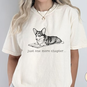 Op de afbeelding: Ivoor kleurig T-shirt met een zwart-wit illustratie van een Corgi die een boek leest. De tekst "just one more chapter..." staat eronder. Het T-shirt heeft korte mouwen en een relaxte pasvorm.