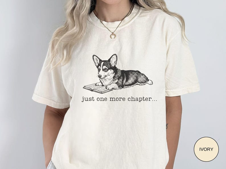 Op de afbeelding: Ivoor kleurig T-shirt met een zwart-wit illustratie van een Corgi die een boek leest. De tekst "just one more chapter..." staat eronder. Het T-shirt heeft korte mouwen en een relaxte pasvorm.