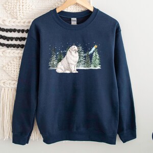 May include: Navy blue sweatshirt featuring a white dog sitting in a snowy forest scene with evergreen trees and a shooting star. The design includes falling snow and a gold star. The sweatshirt is made of a soft material.