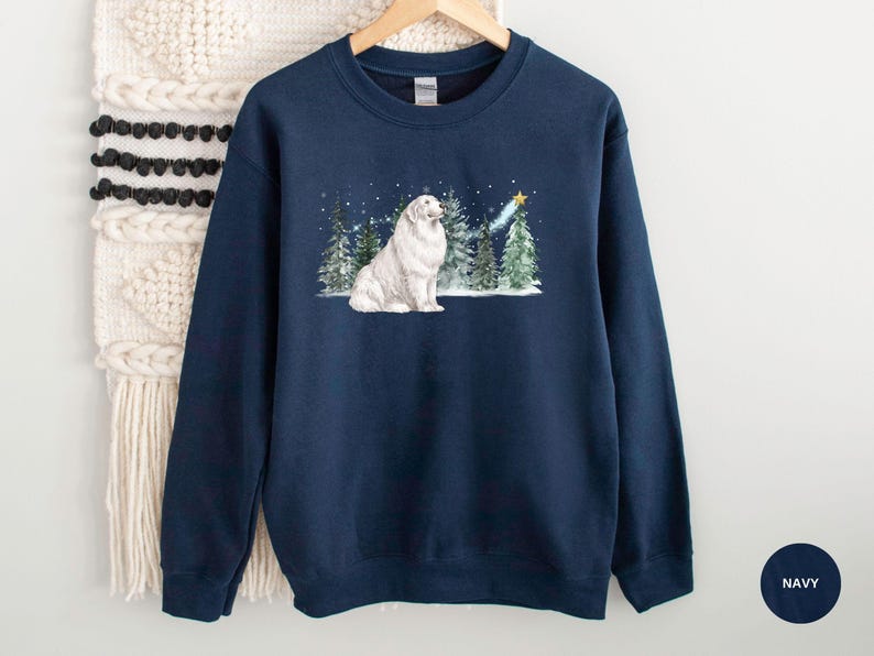 May include: Navy blue sweatshirt featuring a white dog sitting in a snowy forest scene with evergreen trees and a shooting star. The design includes falling snow and a gold star. The sweatshirt is made of a soft material.